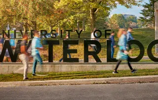 A large sign that reads "UNIVERSITY OF WATERLOO" stands prominently on a grassy area. Blurred figures of people walking by indicate motion, while trees and greenery provide a scenic backdrop.