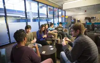 Seven people are sitting in a lounge area with large windows, engaged in a card game. Some are holding cards while others have drinks. Amidst the casual and relaxed atmosphere, a 2nd year student is giving tips near the coffee machine about how to make the most of Reading Week.