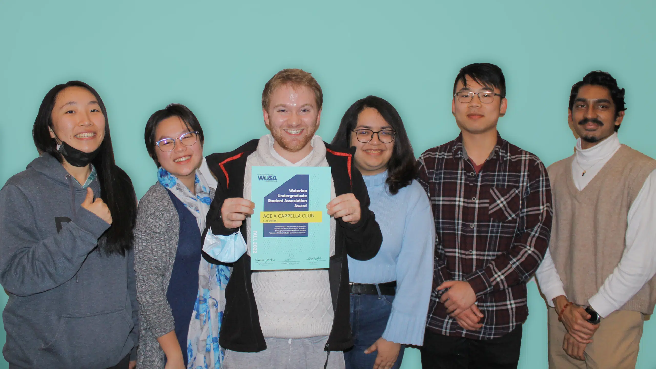 Six people stand in a row, smiling, with one person in the center holding a certificate that reads "WUEA Member, Student Membership, Active Membership to the A Capella Club." They are in casual attire with a light blue background.