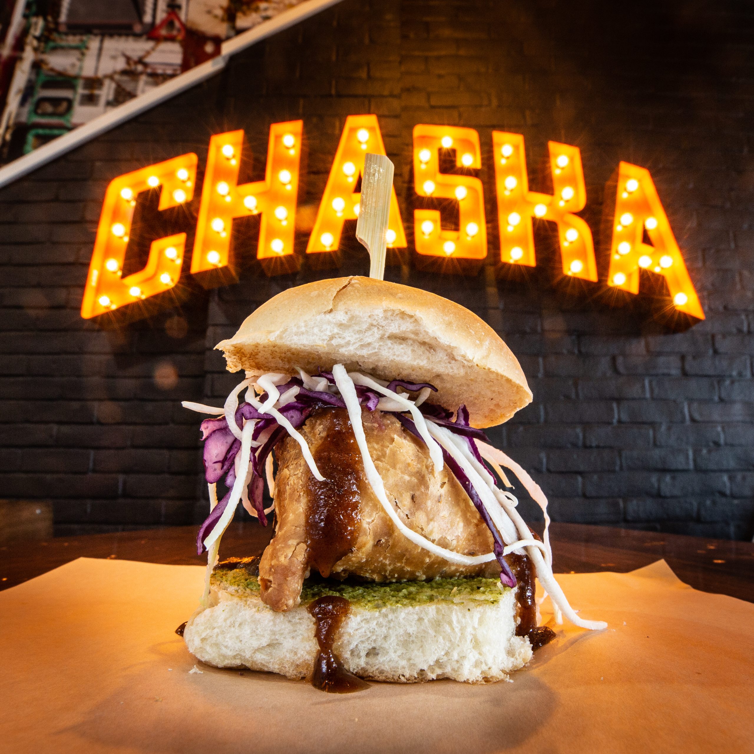 A gourmet burger with shredded cabbage, a crispy fried patty, and sauce is presented on a piece of parchment paper. In the background, an illuminated "CHASKA" sign on a brick wall adds a vibrant touch to the setting.