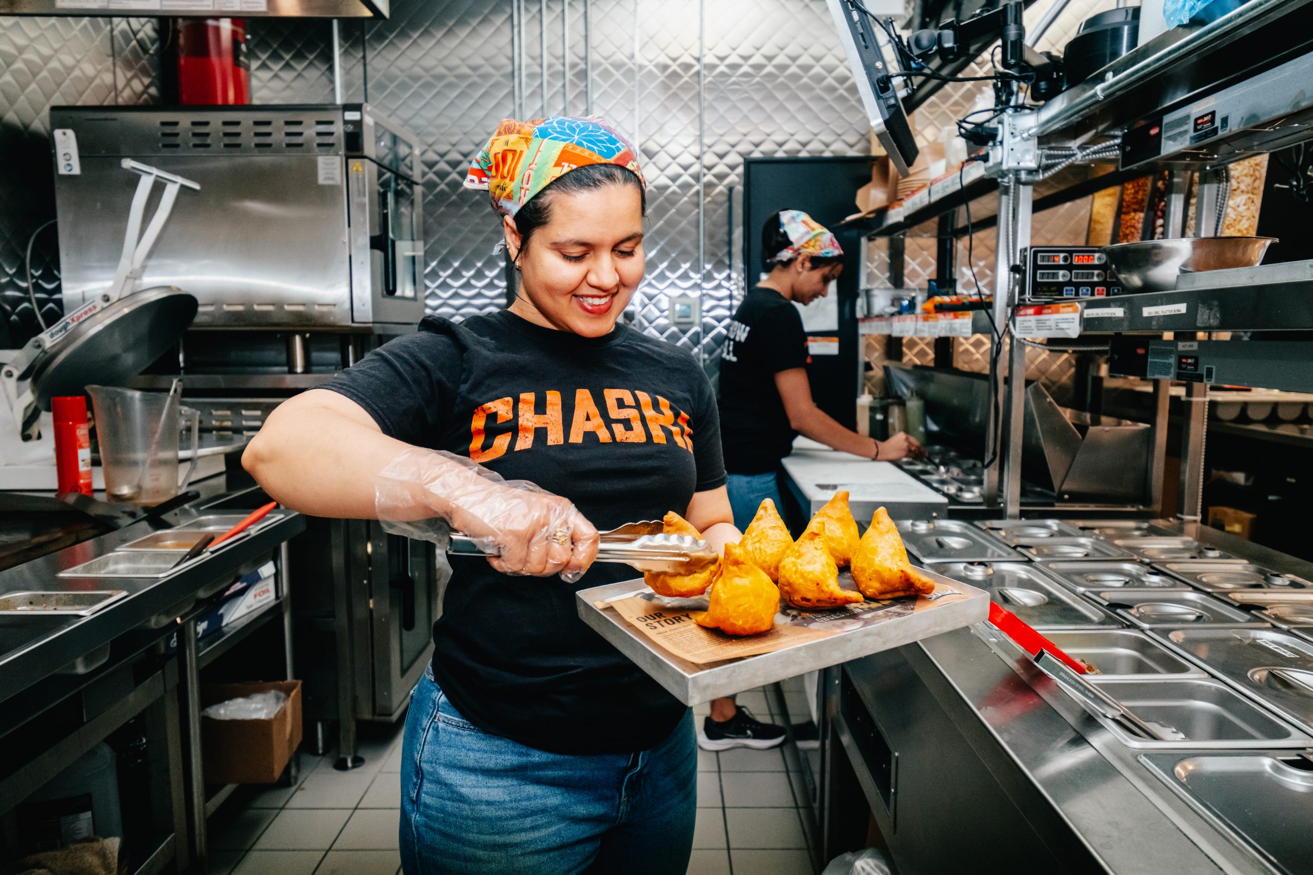 A smiling person wearing a colorful headscarf and a black shirt with the word "CHASKA" is placing samosas on a tray in a commercial kitchen. Another individual can be seen working in the background near a counter with various containers.