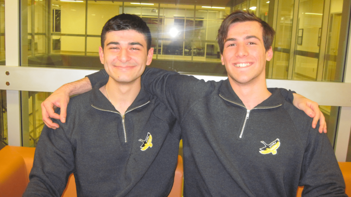 Two young men with short dark hair are sitting side by side on an orange bench. Smiling, they each have an arm around the other's shoulders. Both are wearing matching DDC Quarter Zips featuring a yellow banana logo. The background shows a lit indoor space with windows.