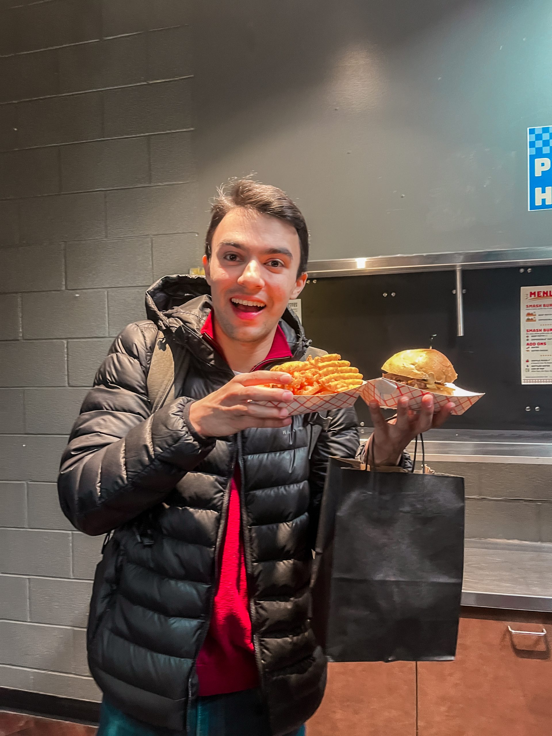 A person with short dark hair, wearing a black puffy jacket and red shirt, smiling and holding a tray of fries and a burger. They also carry a black shopping bag. The background features a dark wall and part of a metal menu holder.