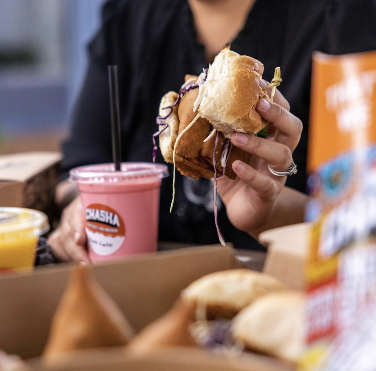 A person holds a sandwich filled with vegetables and sauce in one hand and a pink smoothie with "Chasha" written on the cup in the other. There are various food items and beverages on the table in front of them.