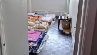 A room with several pillows and blankets arranged on the floor in a makeshift sleeping area, reminiscent of student living at the University of Waterloo. There is a small table at the far end of the room with various items on top and a cardboard box beside it. The wooden floor adds to the sparsely furnished housing landscape.