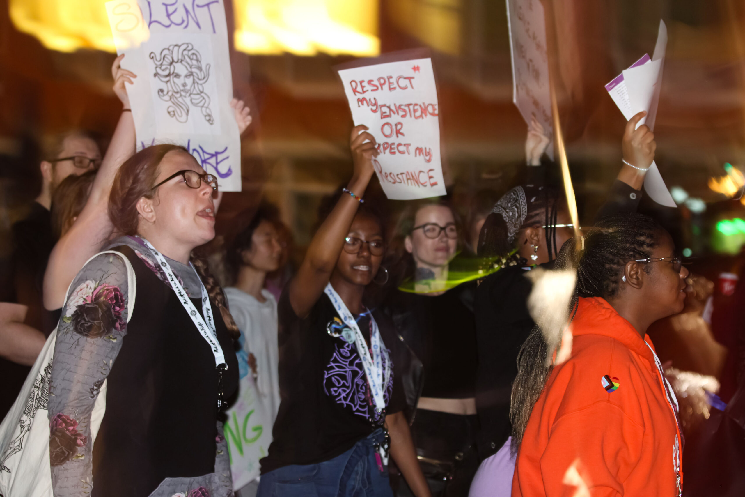 A group of people are seen holding signs and protesting at night during a Take Back the Night event. One protester holds a sign that reads, "Respect my existence or expect my resistance." The scene is energetic, with a diverse group of individuals passionately voicing their message.