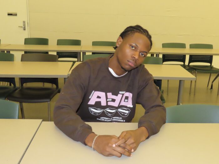 A person with braided hair sits at a desk wearing a TRACES 2024 "Ayo" Sweater in a classroom setting, with empty chairs and tables visible in the background.