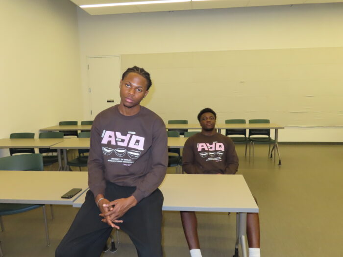 Two people wearing matching TRACES 2024 "Ayo" sweaters sit in an empty classroom, with one sitting on a desk in the foreground and the other sitting at a desk in the background.