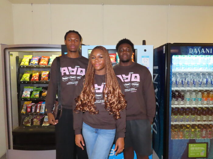 Three individuals stand in front of vending machines, wearing matching dark shirts with a logo that reads "AYO." Sporting their TRACES 2024 "Ayo" Sweater with pride, they examine the machines stocked with snacks and beverages.