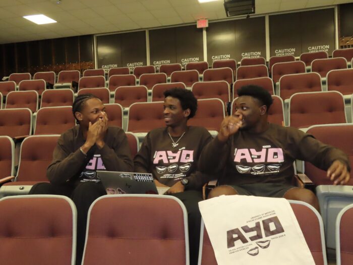 Three individuals wearing matching TRACES 2024 "Ayo" sweaтers are seated in an auditorium, engaged in a lively conversation. Two of them hold open laptops, and a white bag adorned with the "Ayo" logo is on the seat, hinting at their preparation for TRACES 2024.