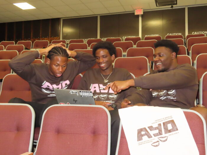 Three men sitting in an auditorium, all wearing matching TRACES 2024 "Ayo" Sweaters, are focused on a laptop. One man holds his head in contemplation while another points at the screen. The third man smiles, embodying the collaborative spirit of TRACES 2024.