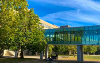 The modern glass walkway between campus buildings glistens under the bright blue sky, surrounded by lush green trees.